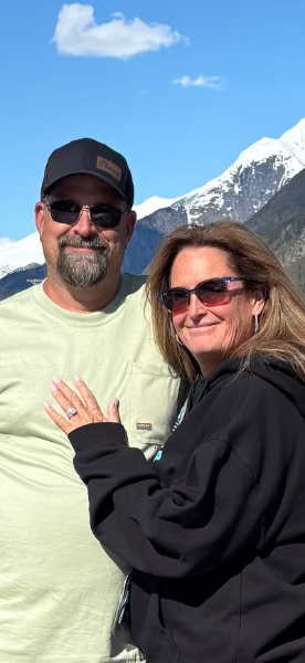 A man and a woman posing outdoors in front of a snow-capped mountain and blue sky. The man wears a black baseball cap, sunglasses, and a light green t-shirt.